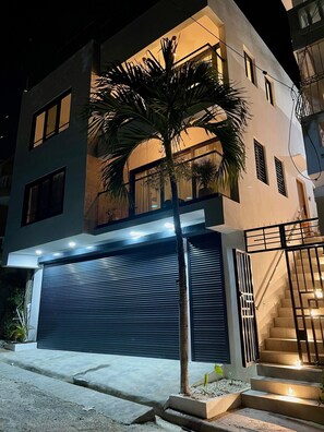A modern two-story building with a garage door, a staircase with lighting, and a palm tree in the foreground. - LEVEL 2, Lovely 2-bedroom apartment with AC steps to malecon in Santa Barbara (Santa Barbara)