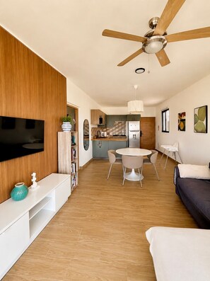 A modern living room with a flat-screen TV, wooden paneling, a ceiling fan, a dining area with a round table and chairs, and a kitchen in the background. - LEVEL 2, Lovely 2-bedroom apartment with AC steps to malecon in Santa Barbara (Santa Barbara)