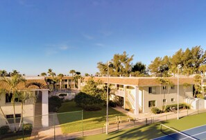 Un hotel de varios pisos con cancha de tenis, palmeras y un cielo azul despejado.