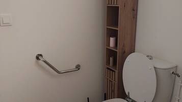 Une salle de bain avec des toilettes blanches, un meuble en bois doté d’étagères, une barre d’appui et un sol carrelé.