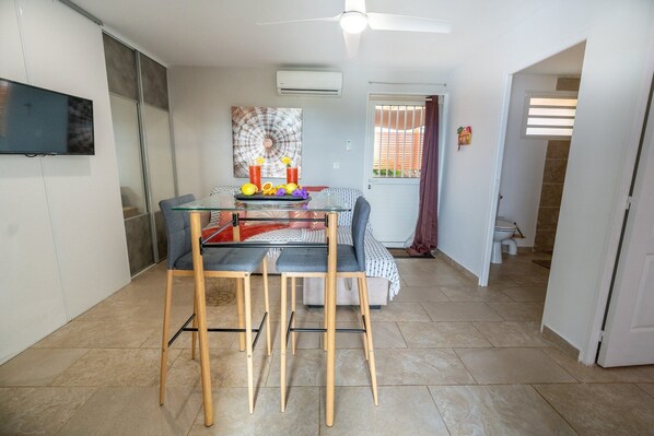 A dining area with a glass table and wooden chairs, a ceiling fan, and a window with blinds.