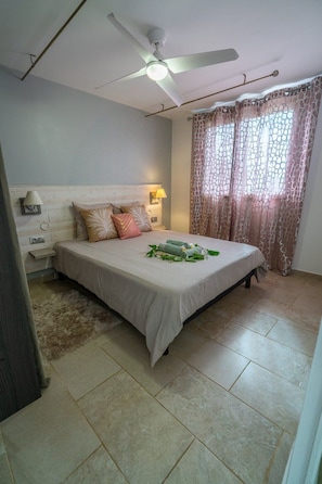 A bedroom with a bed, a ceiling fan, a nightstand, a lamp, and a window with curtains. - Ti Kaz Romance, calm and relaxation (Sainte-Suzanne)