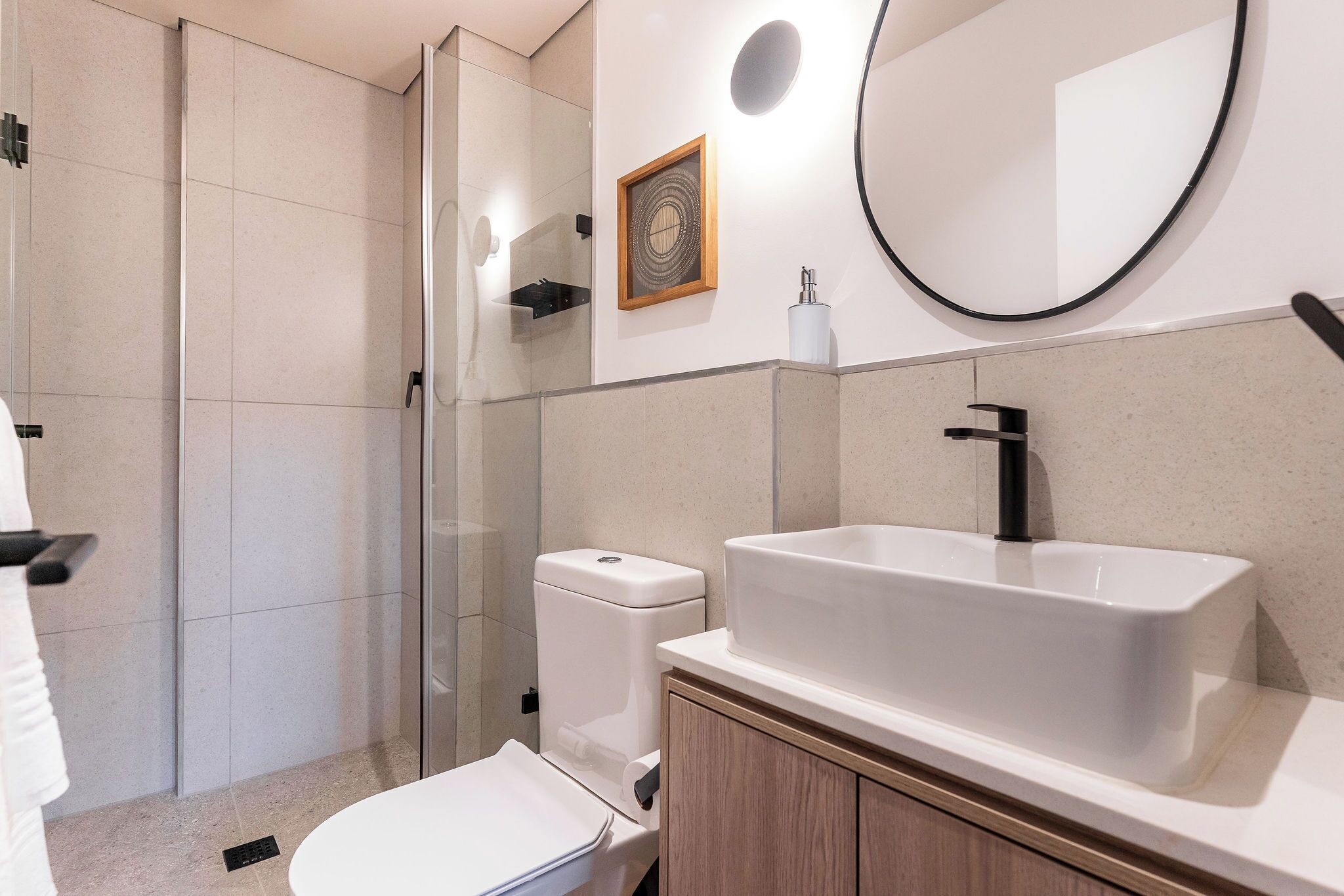 A modern bathroom with a glass shower enclosure, a white toilet, a sink with a black faucet, and a round mirror.