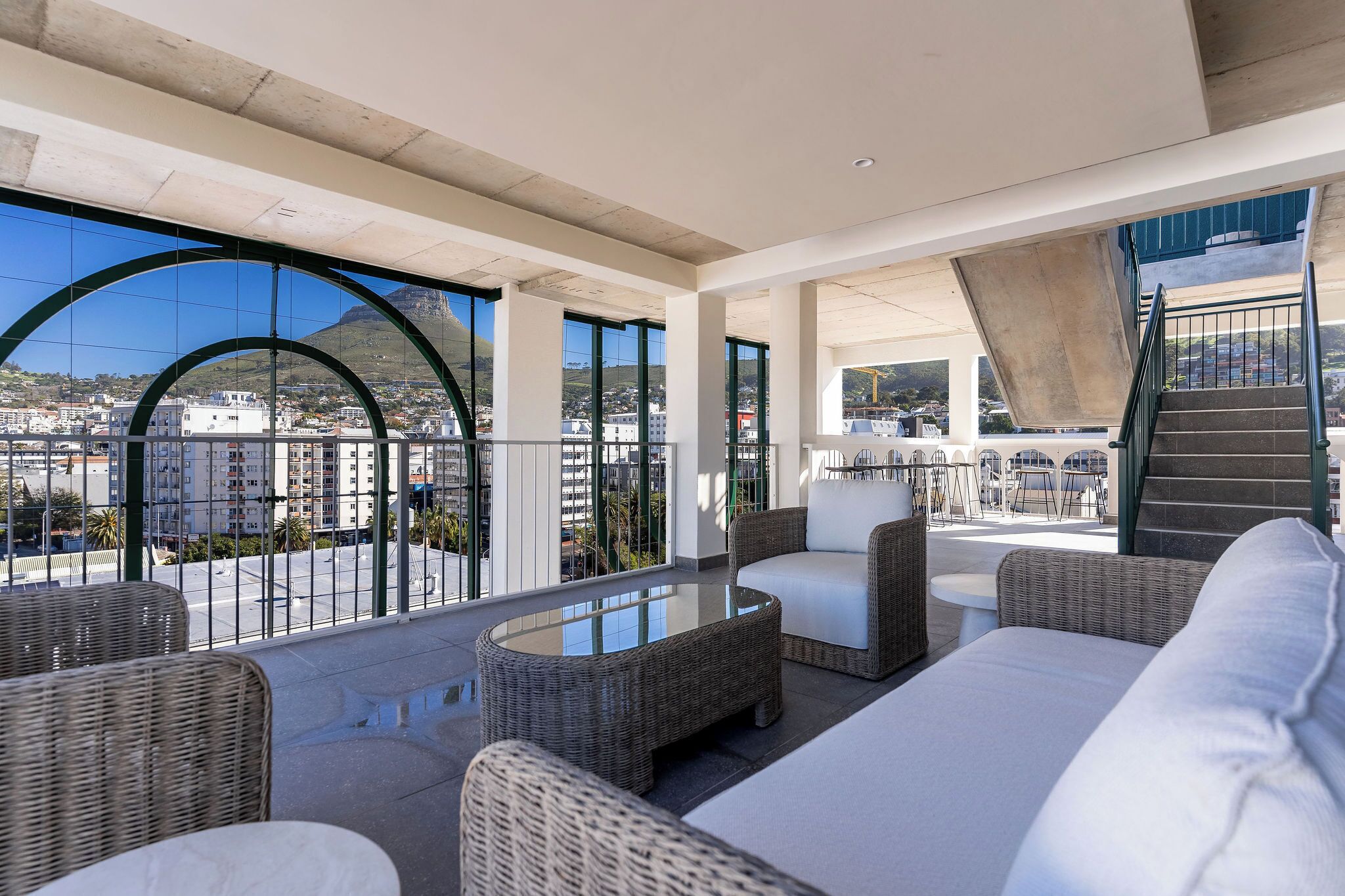 A spacious balcony with wicker furniture and a glass table, overlooking a cityscape with buildings and a mountain.