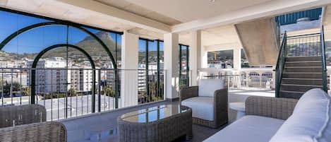 A spacious balcony with wicker furniture and a glass table, overlooking a cityscape with buildings and a mountain.
