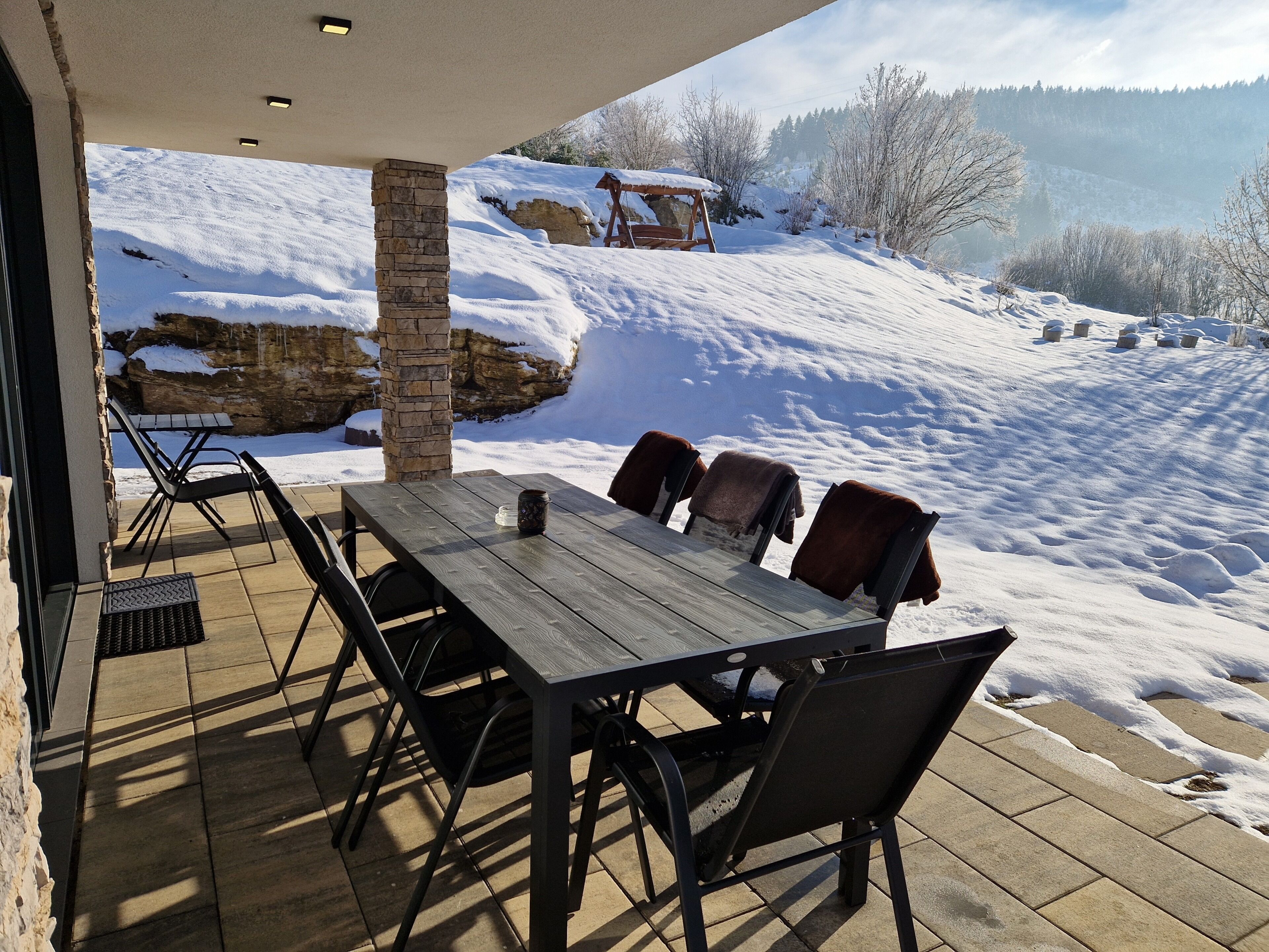 An outdoor patio with a table and chairs, a stone pillar, and a view of a snowy landscape with trees and a rock formation.
