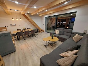 A modern living room with a wooden ceiling, a gray sofa, a dining area with chairs, and a staircase.