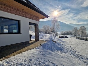 Ein modernes Haus mit einem großen Fenster und einer Steinsäule, umgeben von verschneiter Landschaft und Bergen.
