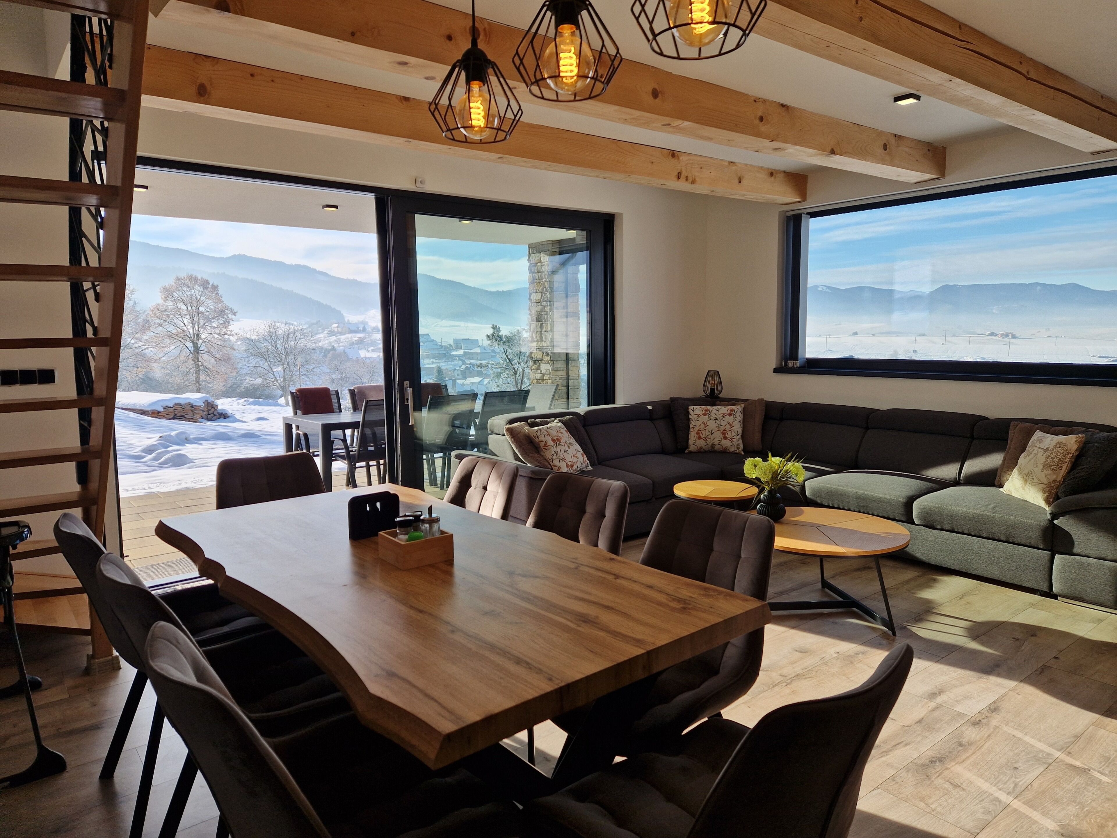 A modern living room with a wooden dining table, comfortable seating, and large windows offering a view of a snowy landscape.