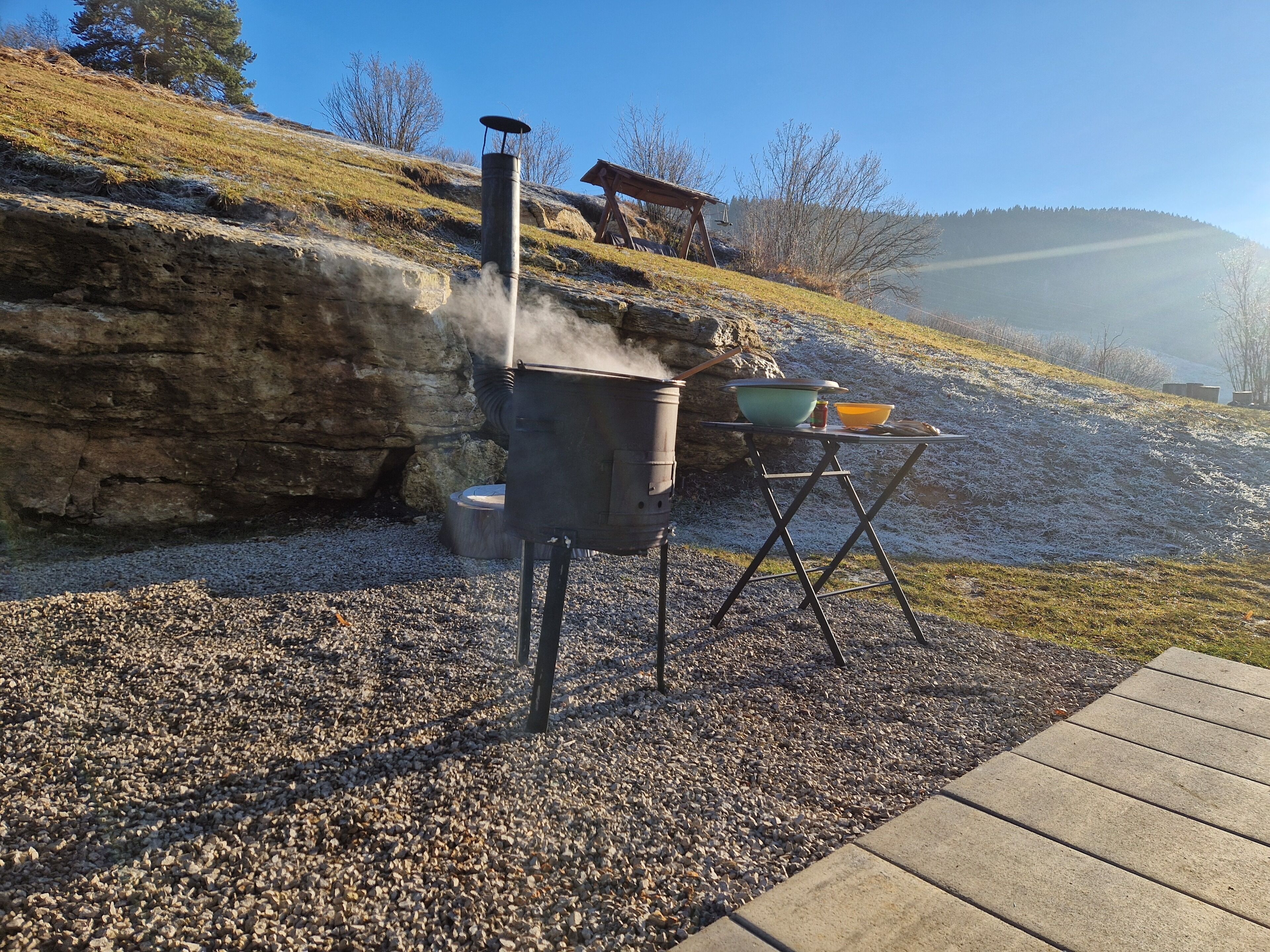 Ett utomhusområde för matlagning med en grill, ett bord med skålar och en stenig terräng.