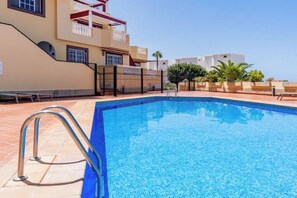 Ein Schwimmbecken mit einer Metallleiter, umgeben von einer gefliesten Terrasse, mit Gebäuden und einem klaren Himmel im Hintergrund.