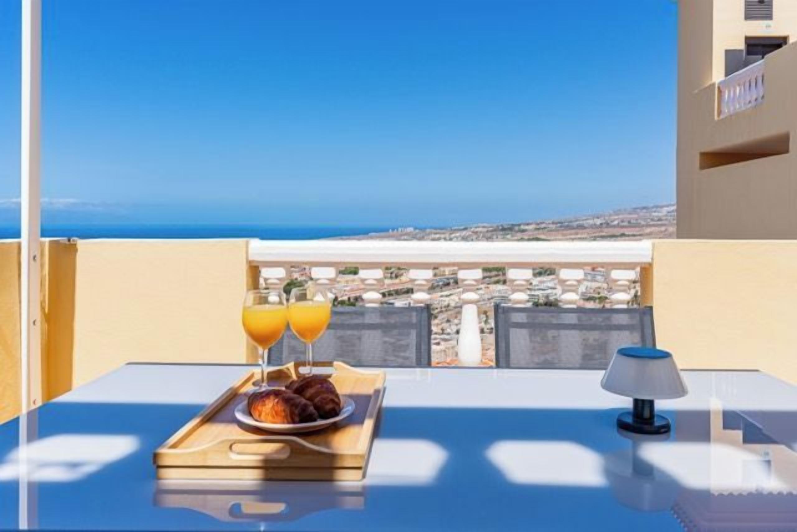 A balcony with a table set for breakfast, a tray with croissants and orange juice, and a view of the city and sea.