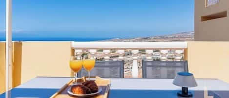 A balcony with a table set for breakfast, a tray with croissants and orange juice, and a view of the city and sea.
