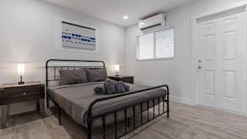 Un dormitorio con cama, mesitas de noche, aire acondicionado de pared, una puerta y una ventana con persianas.