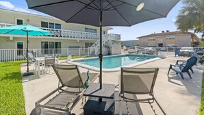Área da piscina com espreguiçadeiras, uma mesa e um grande guarda-sol.