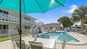 Área de piscina com mesa e cadeiras, um grande guarda-sol e um edifício ao fundo.