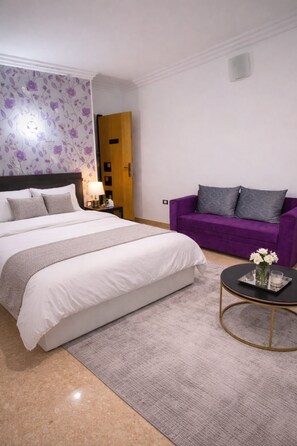 A hotel room with a bed, a purple sofa, a round coffee table, and a wall with a floral pattern. - Family Room in a Luxury Villa (New Cairo)