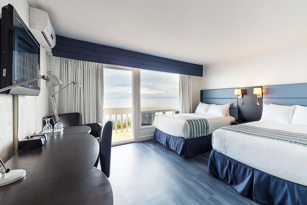 A hotel room with two beds, a desk, a chair, and a large window with a view.