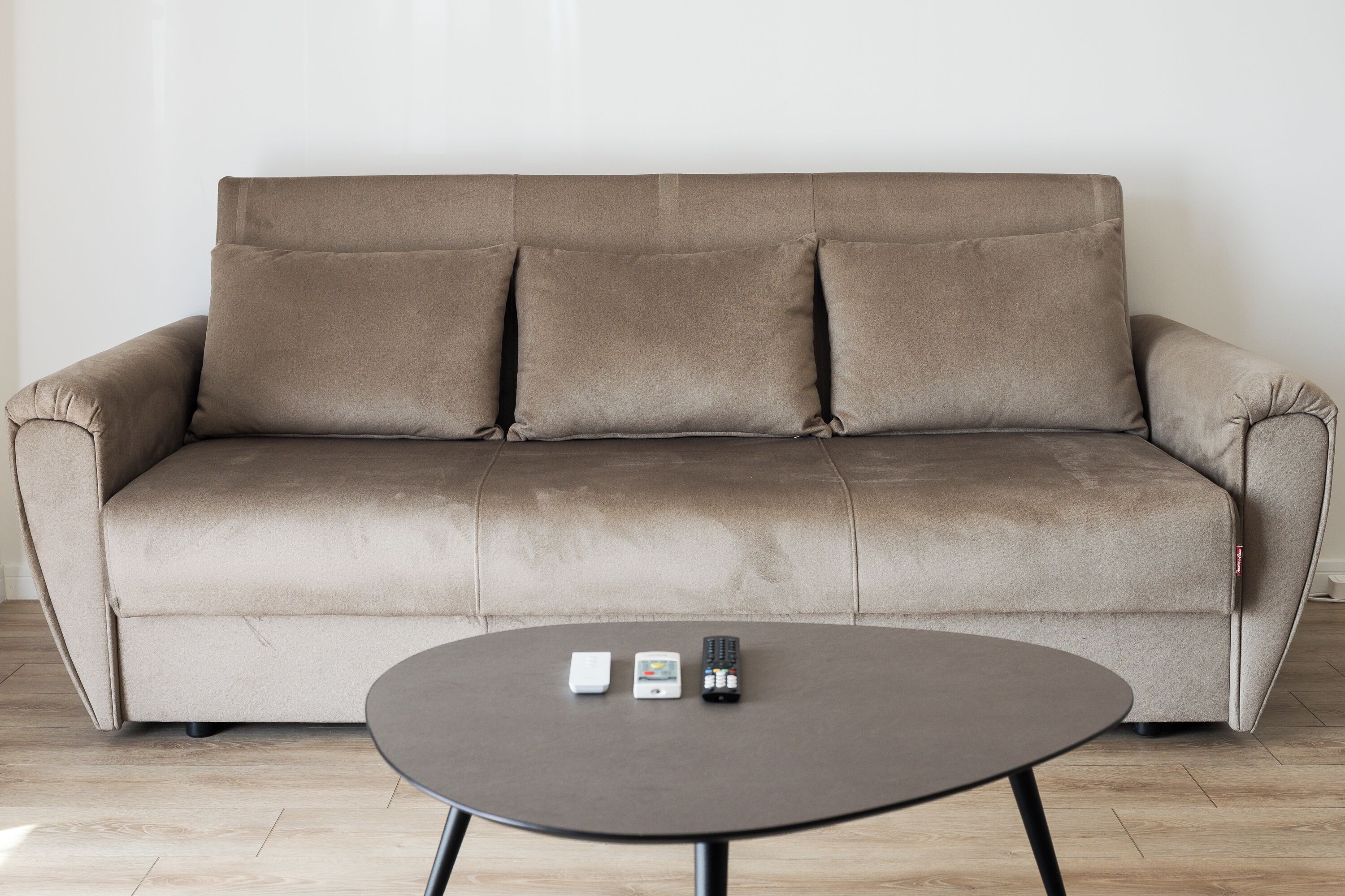 A beige sofa with three cushions and a small round table with two remote controls and a smartphone.
