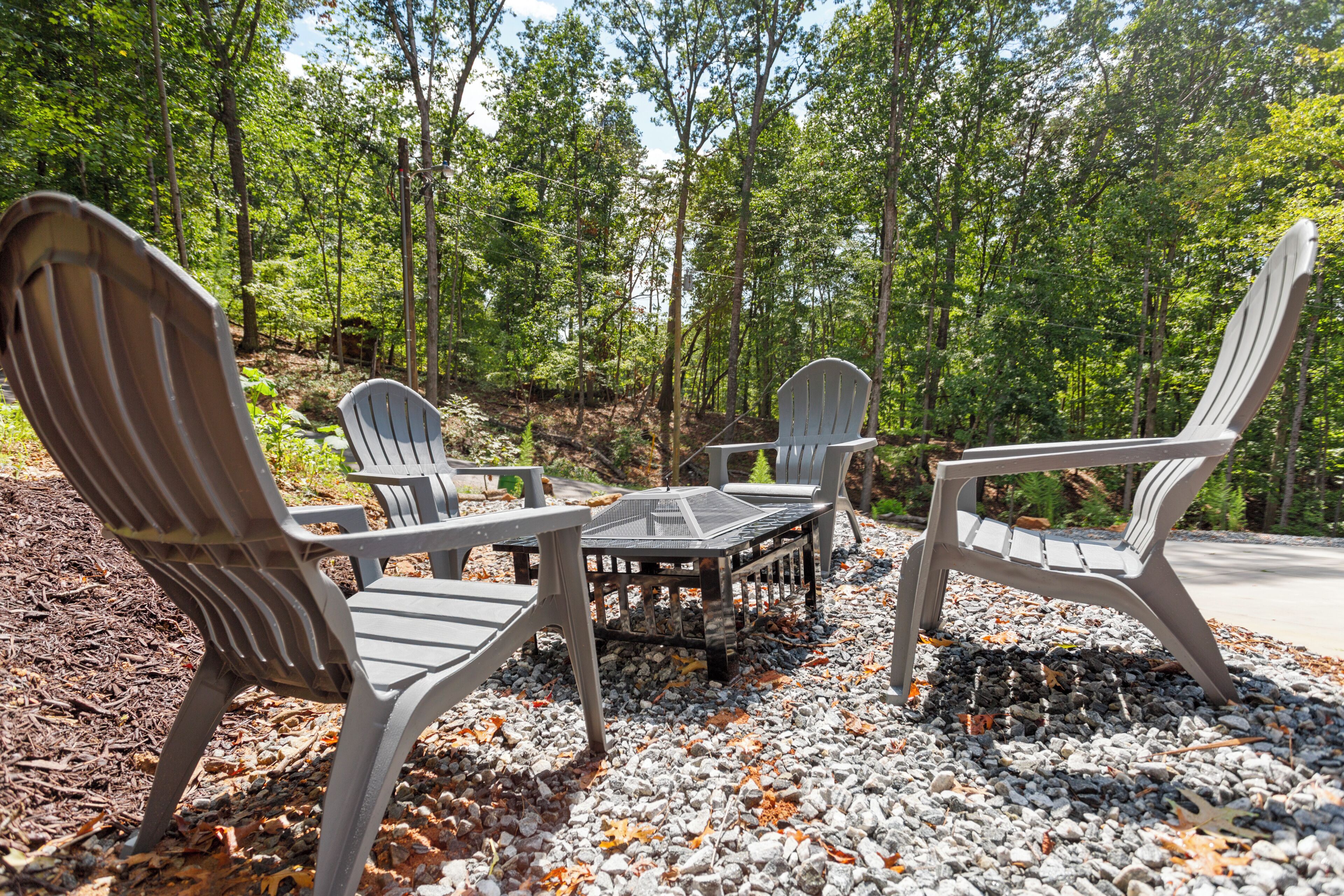 A set of Adirondack chairs and a table in a forested area with gravel ground.