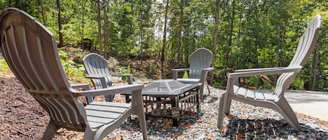 A set of Adirondack chairs and a table in a forested area with gravel ground.