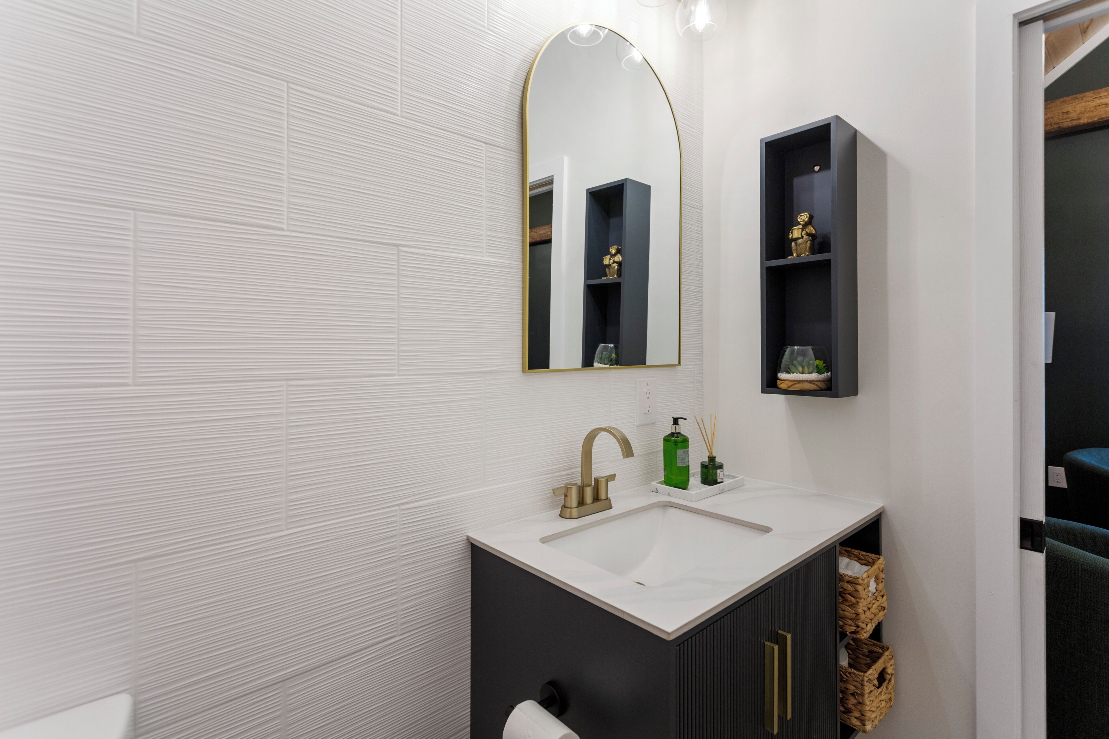 Ein modernes Badezimmer mit einem weißen Waschbecken, einem goldenen Wasserhahn und einem Spiegel mit goldener Rahmung. Über dem Waschbecken befinden sich zwei Regale, eines mit einer goldenen Statue und das andere mit einer grünen Flasche und einer kleinen Pflanze.