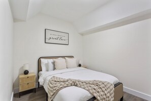 Quarto com cama, mesas de cabeceira, um letreiro fixado na parede e um teto com claraboia.