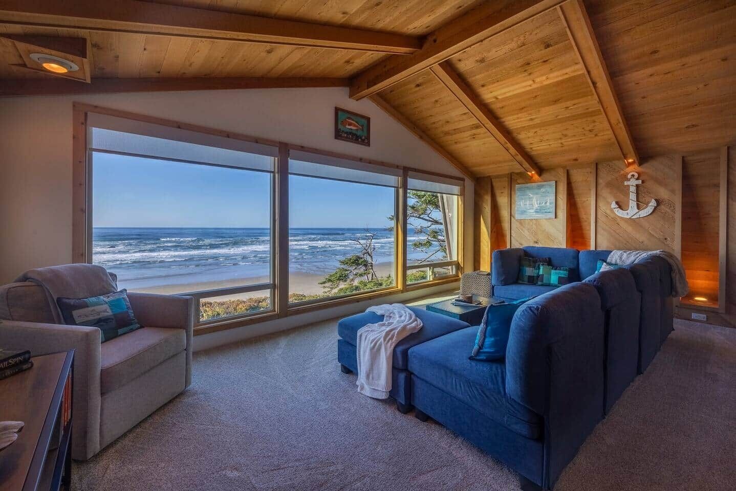 A room with a large window overlooking the ocean, a blue sofa, and a wooden ceiling.