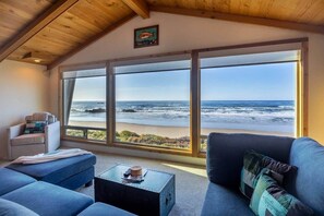 A room with a large window overlooking the ocean, a blue sofa, a small table with a basket, and a chair. - Solitude in Seal Rock (Seal Rock)