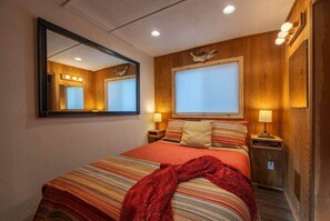 Une chambre d’hôtel comprenant un lit, un miroir, deux lampes de chevet, une fenêtre avec des stores et un panneau mural en bois.