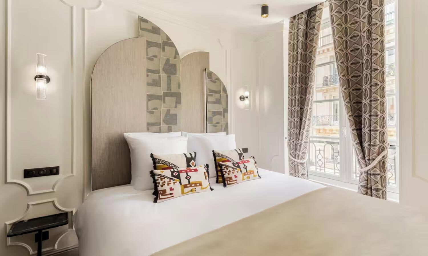 Une chambre d’hôtel avec un grand lit, des coussins décoratifs, un mur orné d’un motif géométrique et une fenêtre avec des rideaux à motifs.