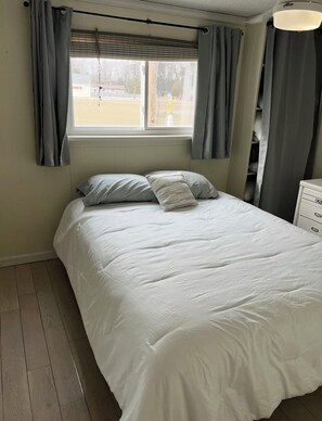 A bedroom with a bed, grey curtains, a window, and a bedside table. - Waterfront Escape - Bay View Beach House (Tawas City)