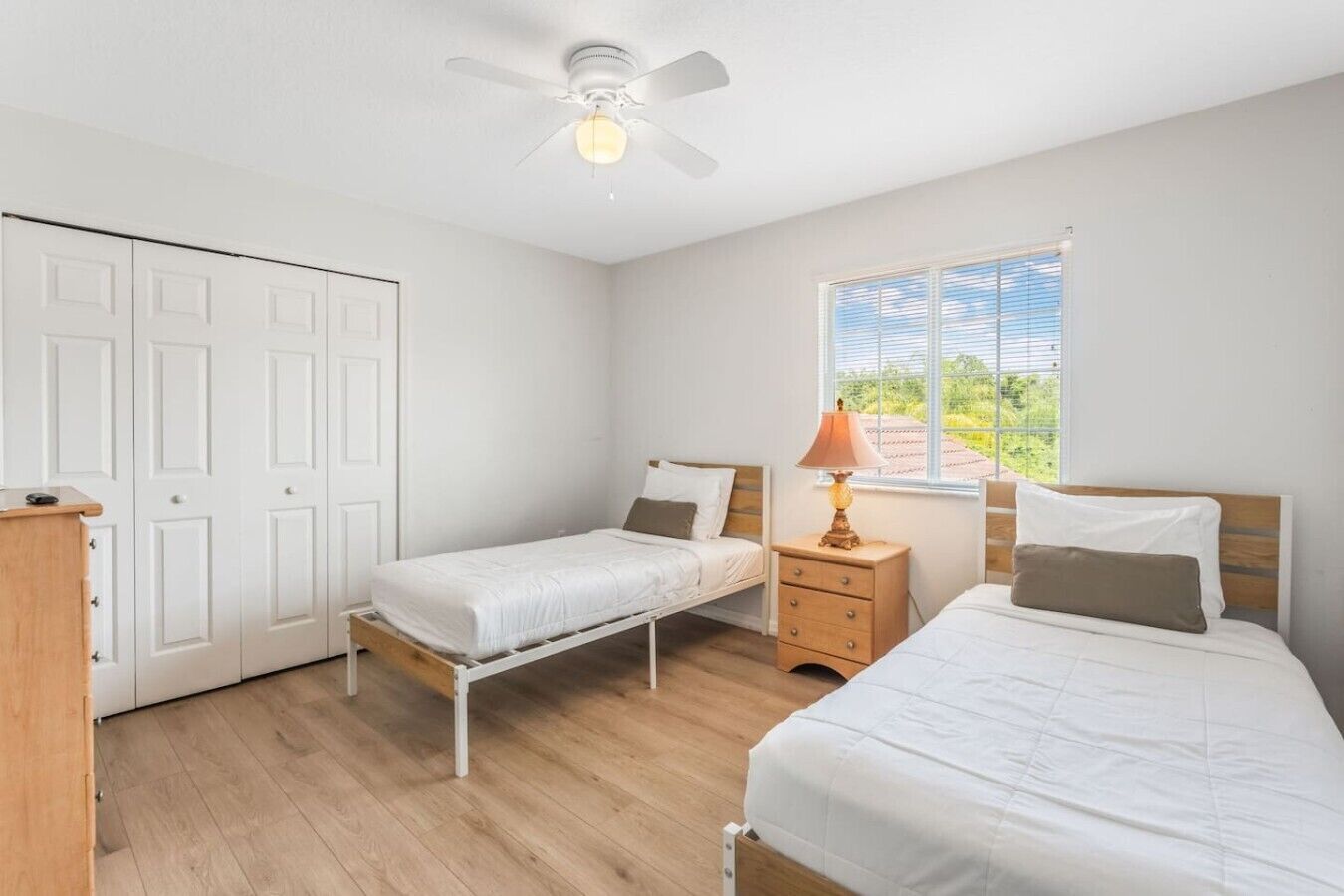 A bedroom with two single beds, a ceiling fan, a window with blinds, and a bedside table with a lamp.