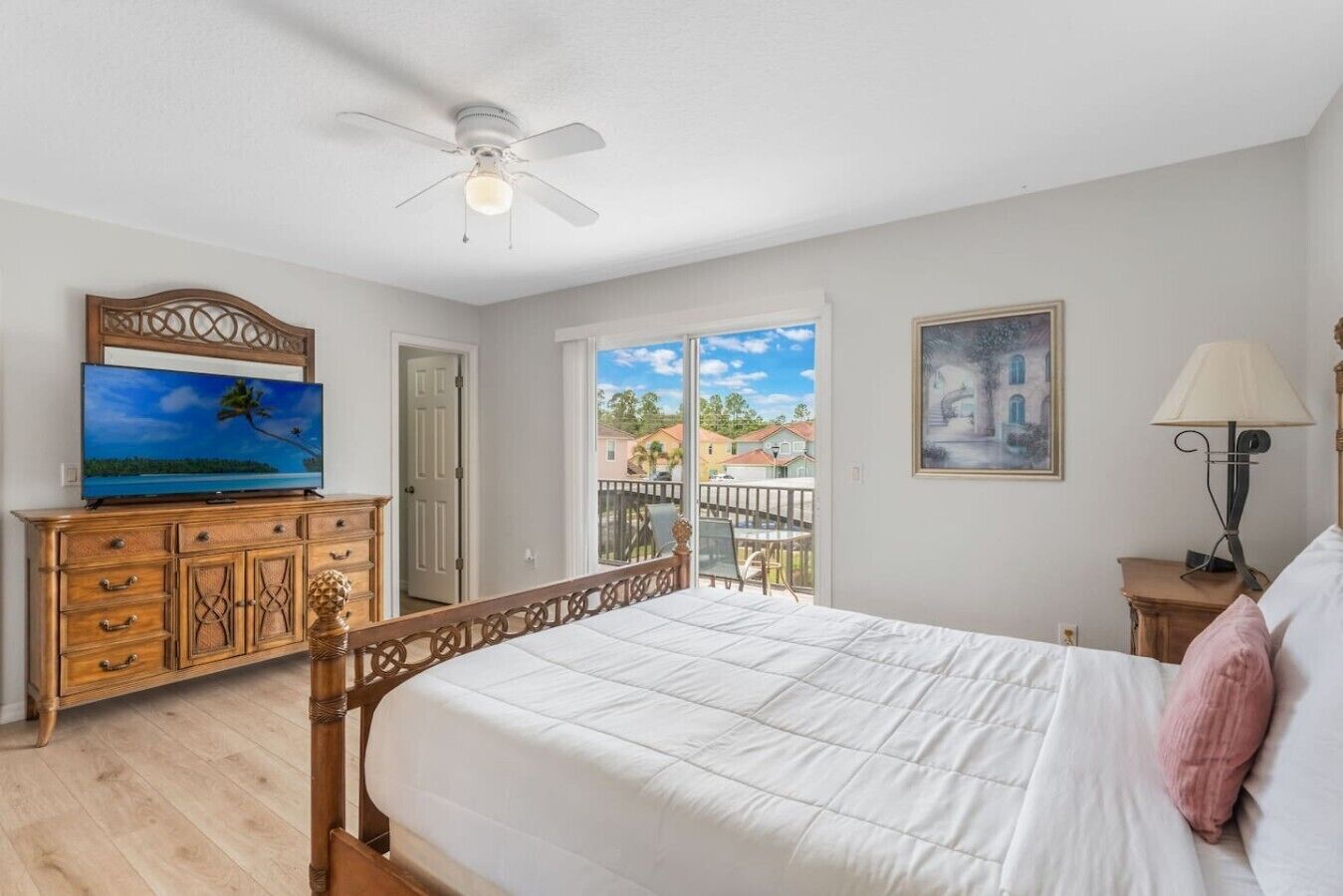 Une chambre à coucher avec un lit, un téléviseur posé sur une commode en bois, un ventilateur de plafond, un balcon avec vue et un tableau accroché au mur.
