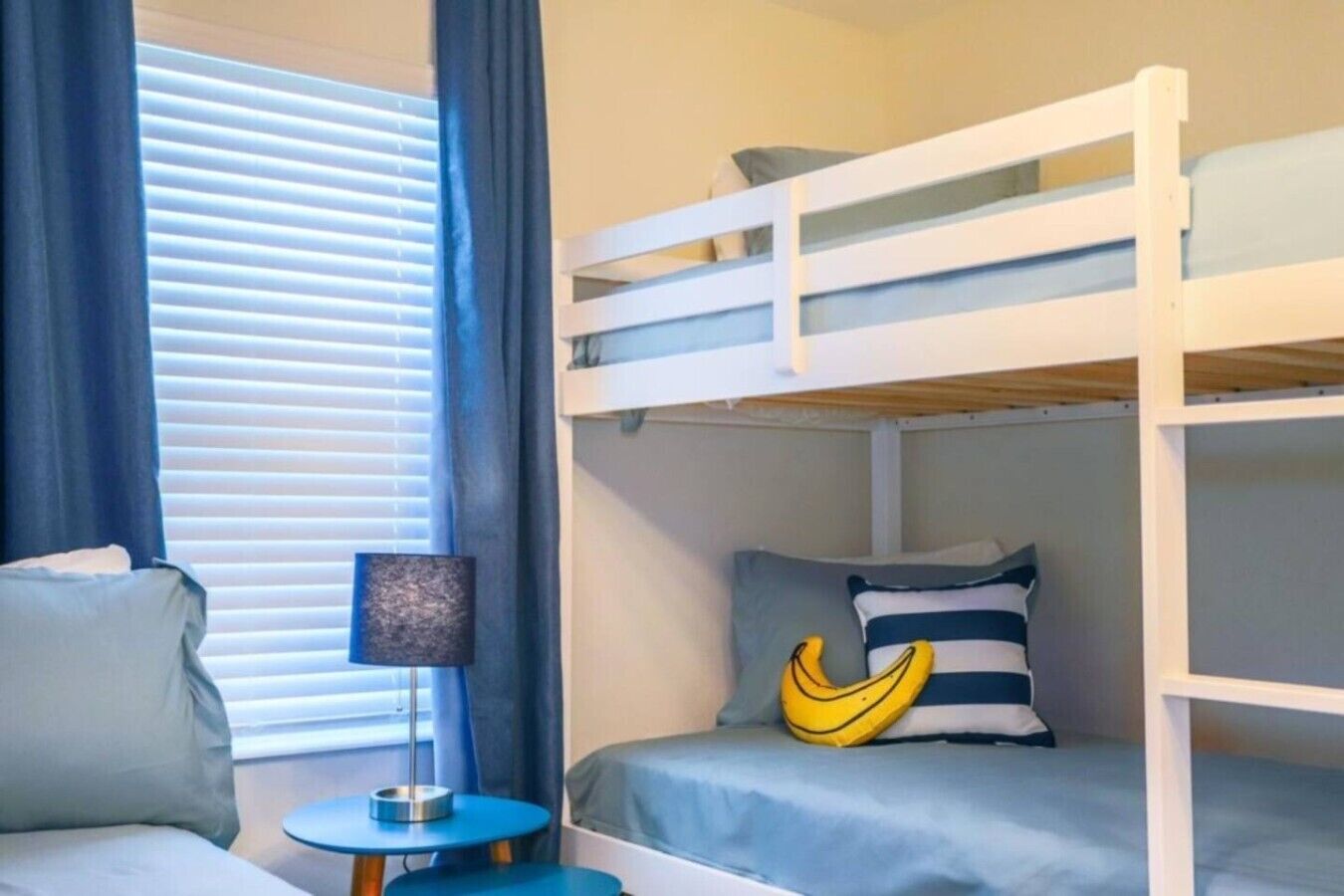 A bunk bed with a pillow and a banana-shaped pillow, a small round table with a lamp, and a window with blinds.