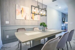 Un espace de salle à manger moderne avec une longue table, six chaises, des œuvres d’art abstraites accrochées au mur et un luminaire suspendu.