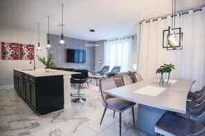 A modern kitchen with a central island, a dining area with a table and chairs, and a living space with a TV and seating. - Themed Championsgate private pool water park BBQ (Kissimmee)