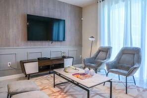 A modern living room with a flat-screen TV, two grey armchairs, a coffee table with books, and a patterned rug. - Themed Championsgate private pool water park BBQ (Kissimmee)