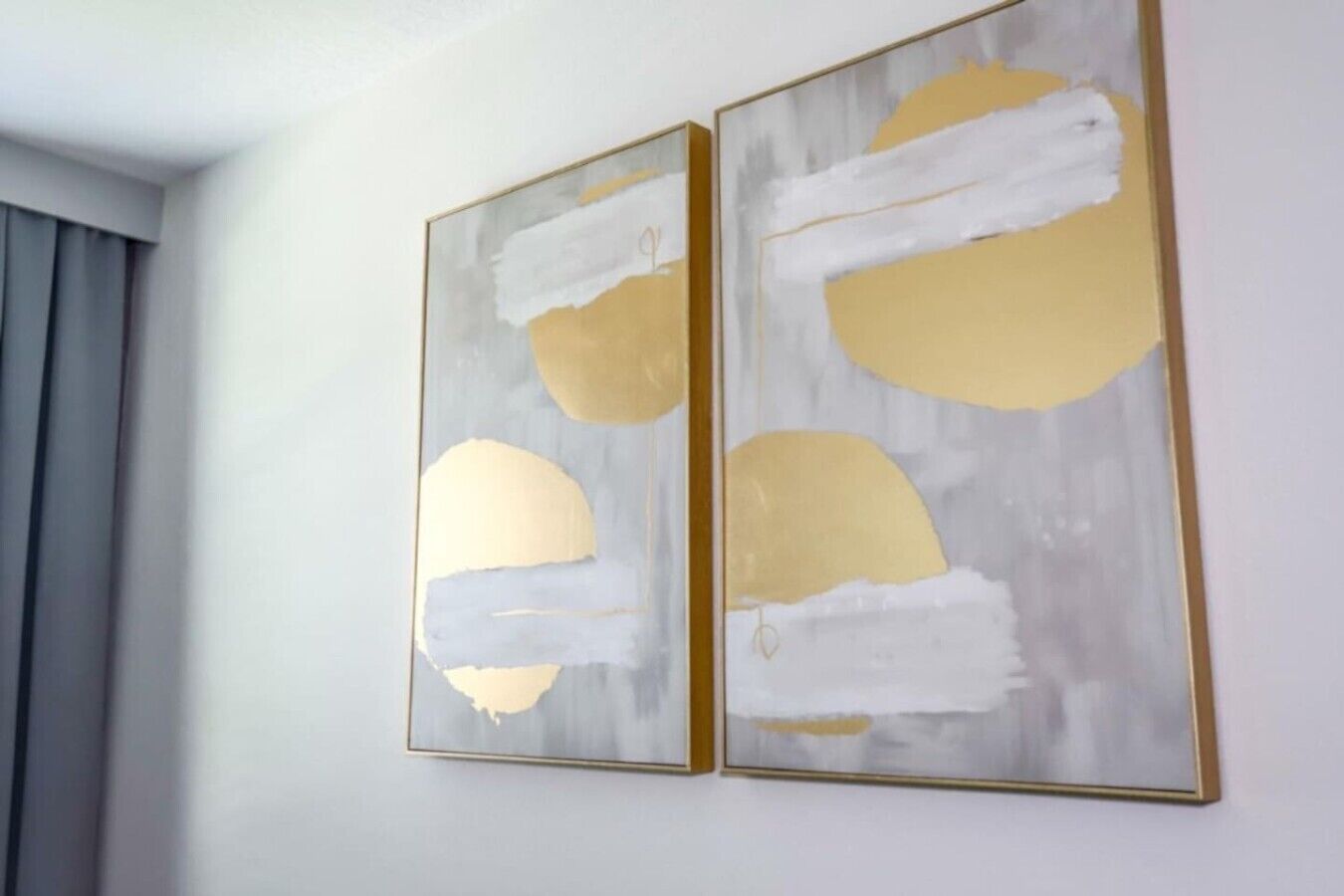 Zwei abstrakte Gemälde mit goldenen und weißen Pinselstrichen an einer hellen Wand.