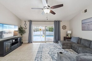 Ein Wohnzimmer mit Deckenventilator, Flachbildfernseher, grauem Sofa, gemustertem Teppich und einer Glastür, die zu einem Pool führt.