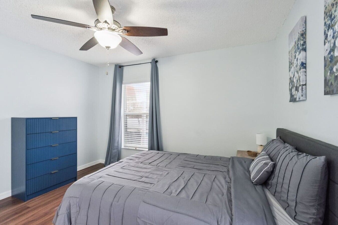 Une chambre à coucher avec un lit, un ventilateur de plafond, une fenêtre avec des rideaux et une commode bleue.