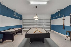 A pool table with a cue stick and a billiards table in a room with blue walls and a garage door in the background. - Bella Blue at Bella Vida Resort (Kissimmee)