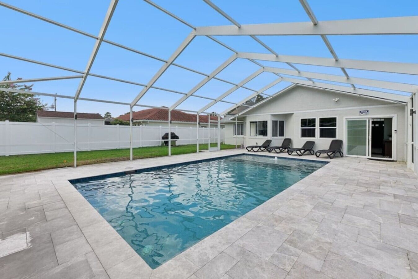 A covered swimming pool with a clear blue water surface, surrounded by a white fence, and a building with a door and windows in the background.
