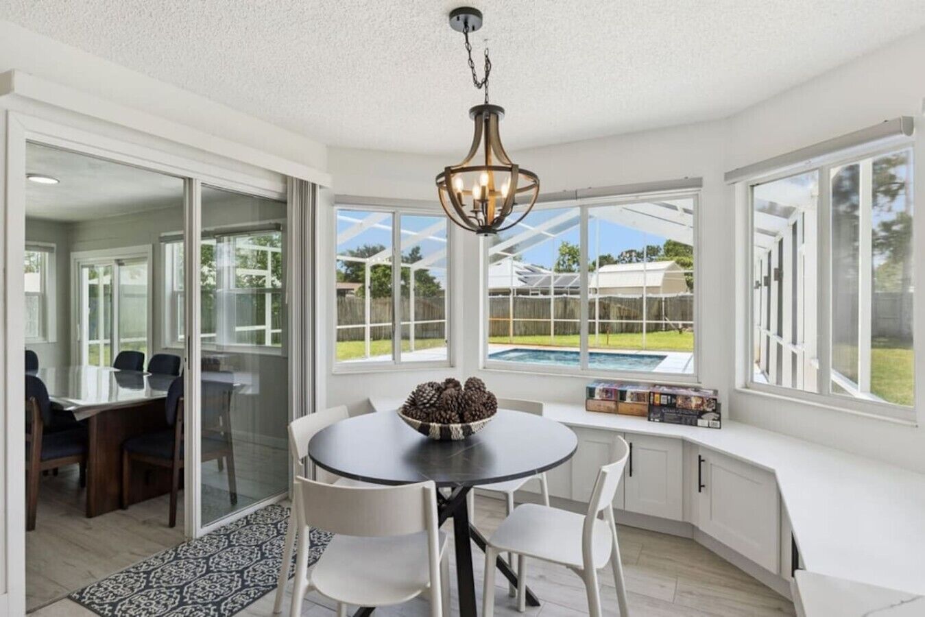 A sunlit room with a dining table, chairs, a chandelier, and a view of a swimming pool.