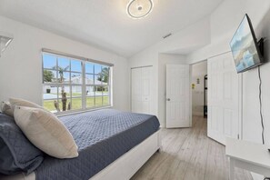 Un dormitori amb un llit, una finestra, un televisor i una porta que dona a una altra habitació.