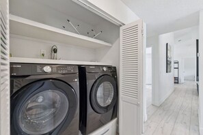 Una sala de bugaderia amb dues rentadores Samsung negres, una estant blanca amb una planta i una porta blanca amb persianes.