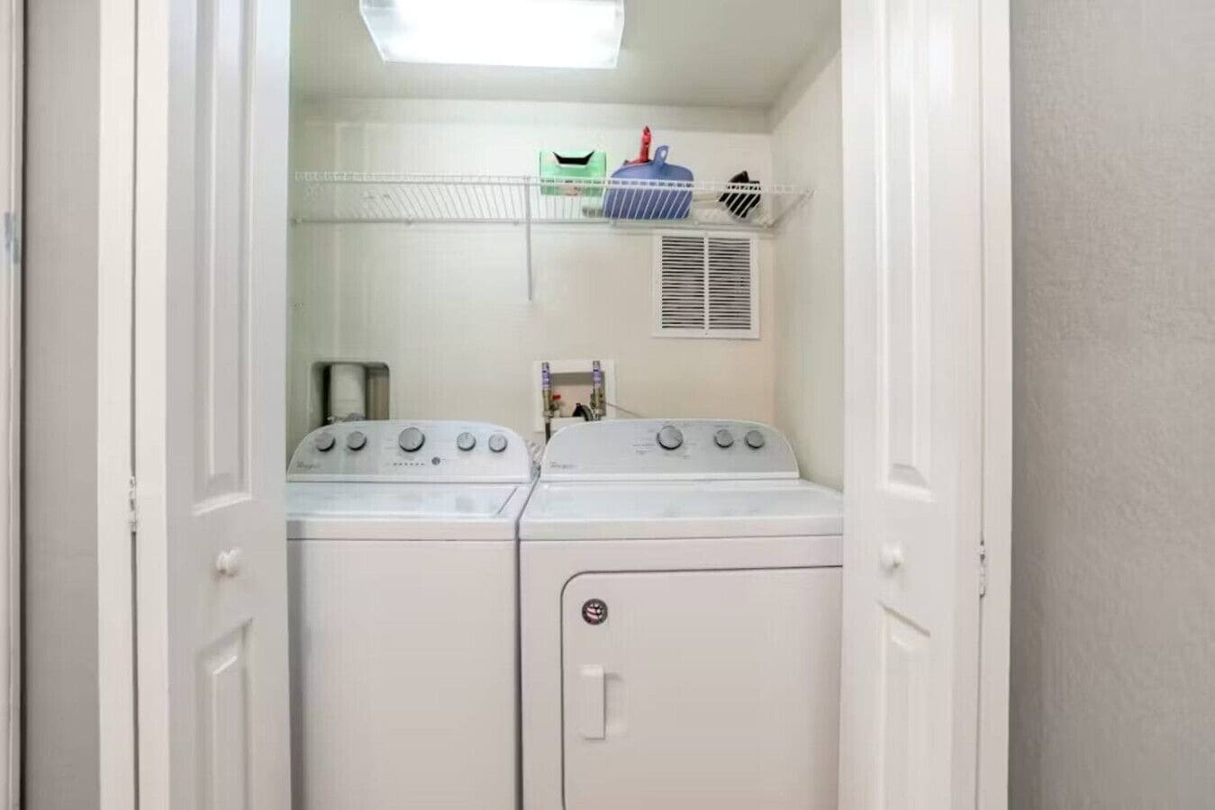 A laundry room with a front-loading washer and dryer, a shelf with laundry supplies, and a vent above the dryer.