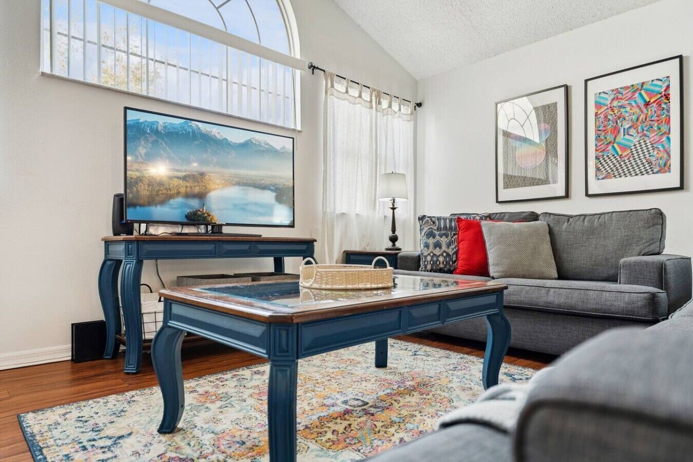 Ein Wohnzimmer mit einem großen Flachbildfernseher, einem blauen Couchtisch, einem grauen Sofa und einem farbenfrohen Teppich.