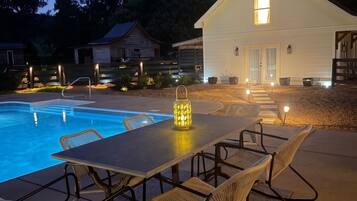 Un espace aménagé au bord de la piscine avec une table et des chaises, une petite maison en arrière-plan et un ciel dégagé.