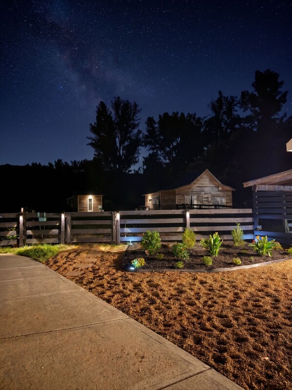 A starry night sky with a wooden cabin and a fenced garden area.